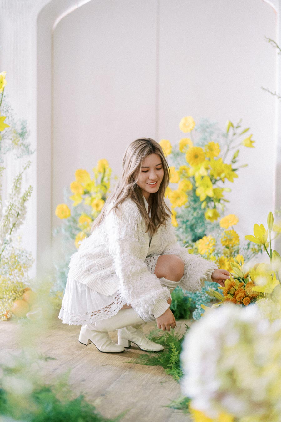 Young woman in a cozy white outfit kneeling among vibrant yellow flowers and greenery, creating a fresh and cheerful bridal or spring theme in a light-filled room.