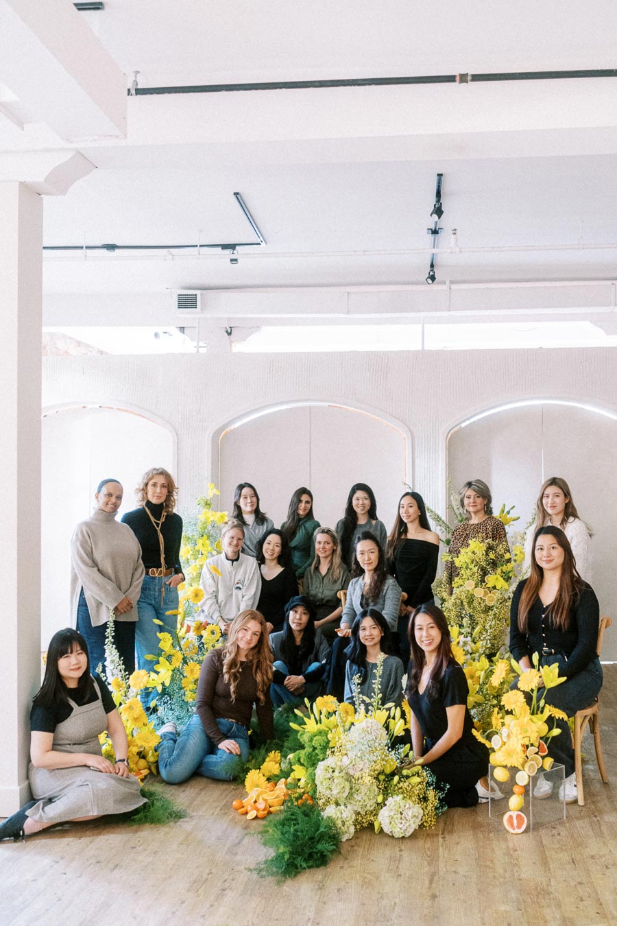 A group of people posing together in a bright room decorated with vibrant yellow flowers and greenery. The scene conveys a sense of teamwork and creativity.