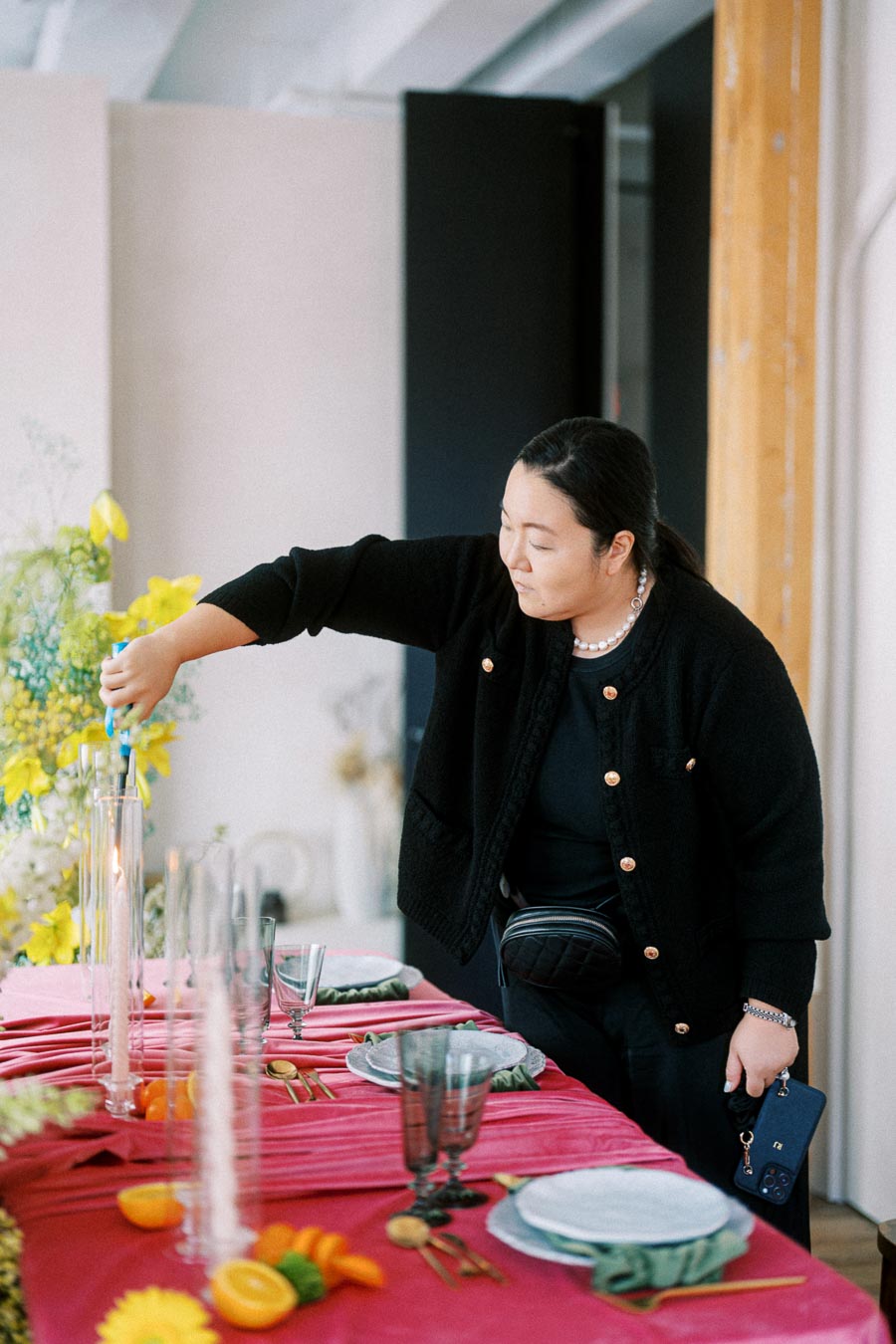 A woman in a black outfit lights a candle on a beautifully set table with pink tablecloth, adorned with orange slices and green plants, preparing for an elegant event.