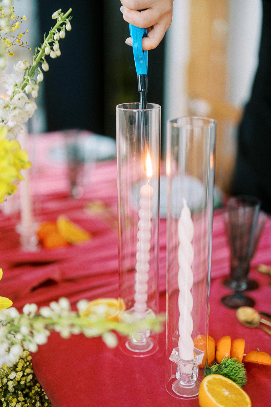 A person uses a blue lighter to ignite a spiral white candle in a tall glass holder on a vibrant pink tablecloth, adorned with fresh flowers and citrus fruit slices, creating an elegant and festive table setting.