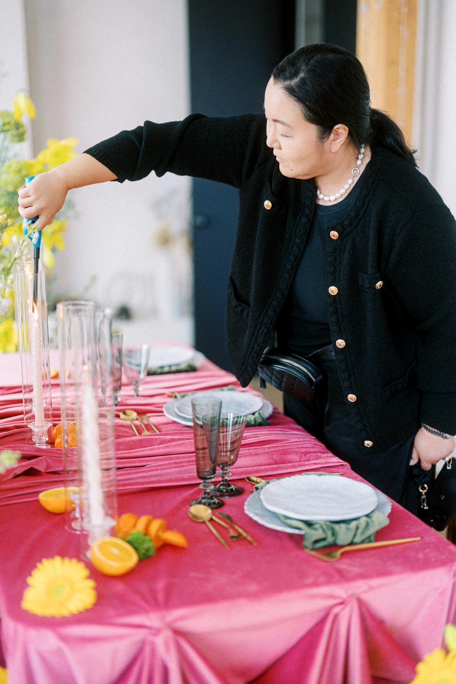 A person lighting a candle on a stylishly set dining table with a pink tablecloth, elegant glassware, gold cutlery, and decorative orange slices and flowers, creating a vibrant and inviting atmosphere.