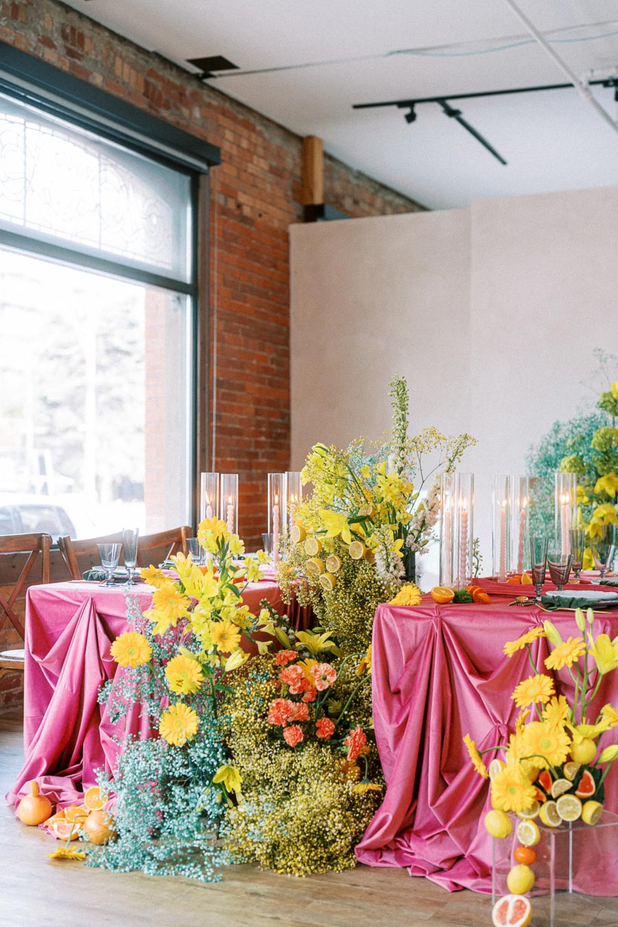 Luxurious event table setting with vibrant pink tablecloth and an elaborate floral arrangement featuring yellow and orange blooms, accented with sliced citrus fruits and tall candles, set against a rustic brick wall.