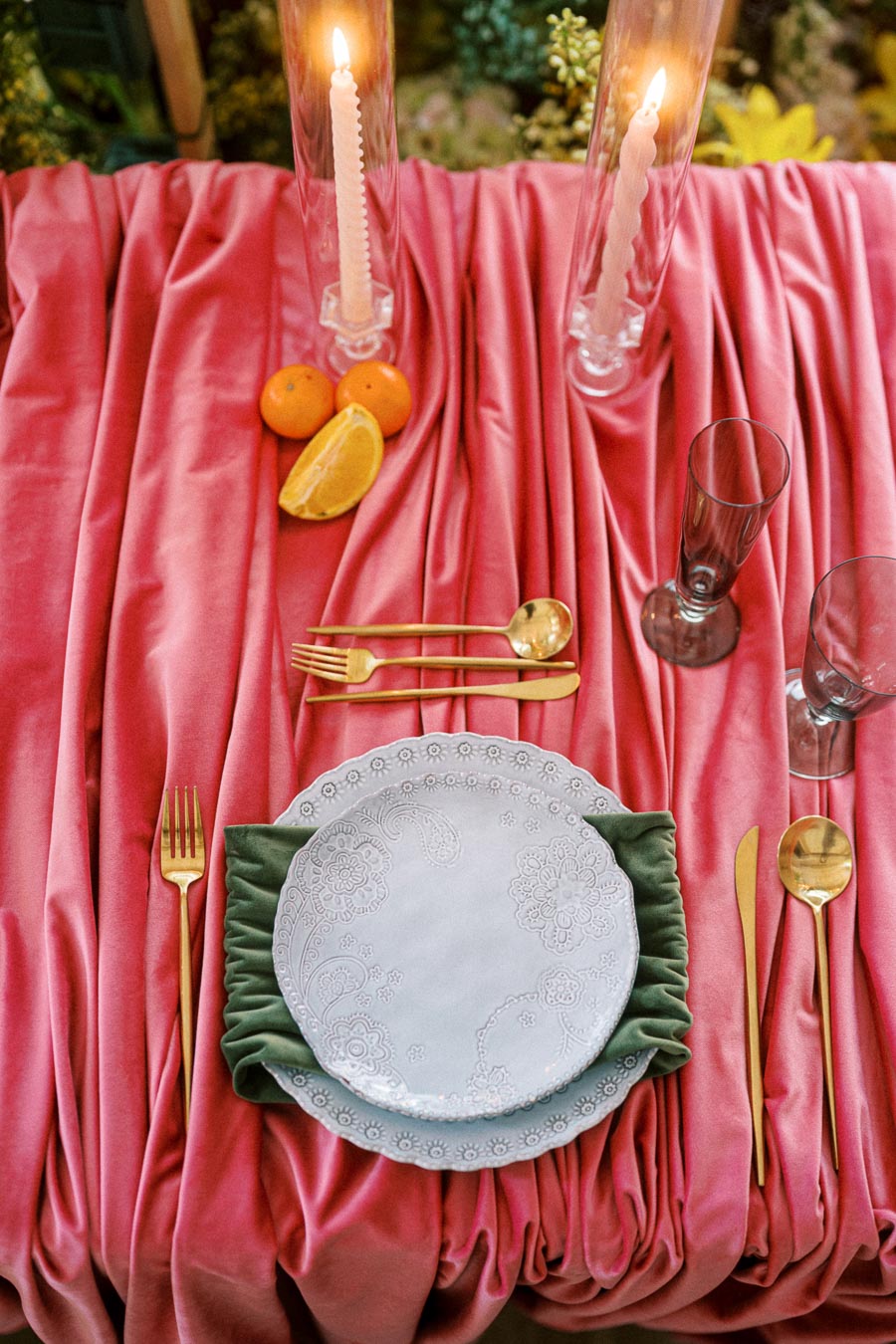Luxurious tablescape with a pink velvet tablecloth, elegant white dinnerware with floral patterns, and gold cutlery, set alongside a green napkin. Two glowing candles and fresh citrus fruits complete the sophisticated dining setting.