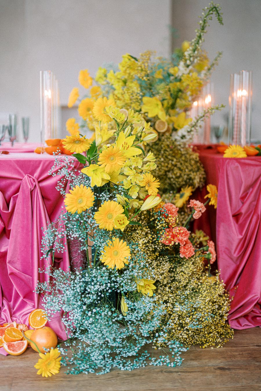 Lush floral arrangement with vibrant yellow and orange flowers, accompanied by green foliage and sliced oranges, set against a table with rich pink drapery and lit candles for an elegant event setting.