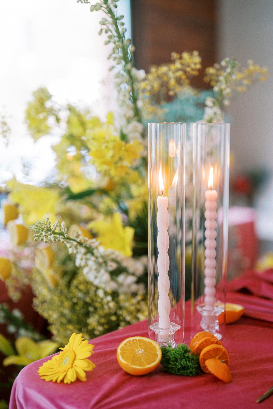Elegant table setting featuring tall, twisted candles in glass holders surrounded by vibrant yellow flowers and fresh orange slices on a pink tablecloth.