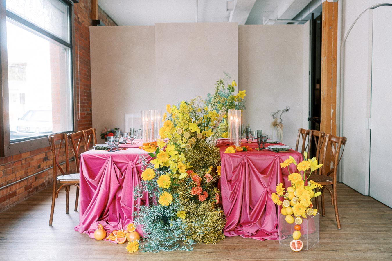 Elegant indoor wedding table setting with vibrant pink tablecloths, adorned with abundant yellow floral arrangements and citrus decor, surrounded by wooden chairs, and set near a large window in a stylish brick-walled venue.
