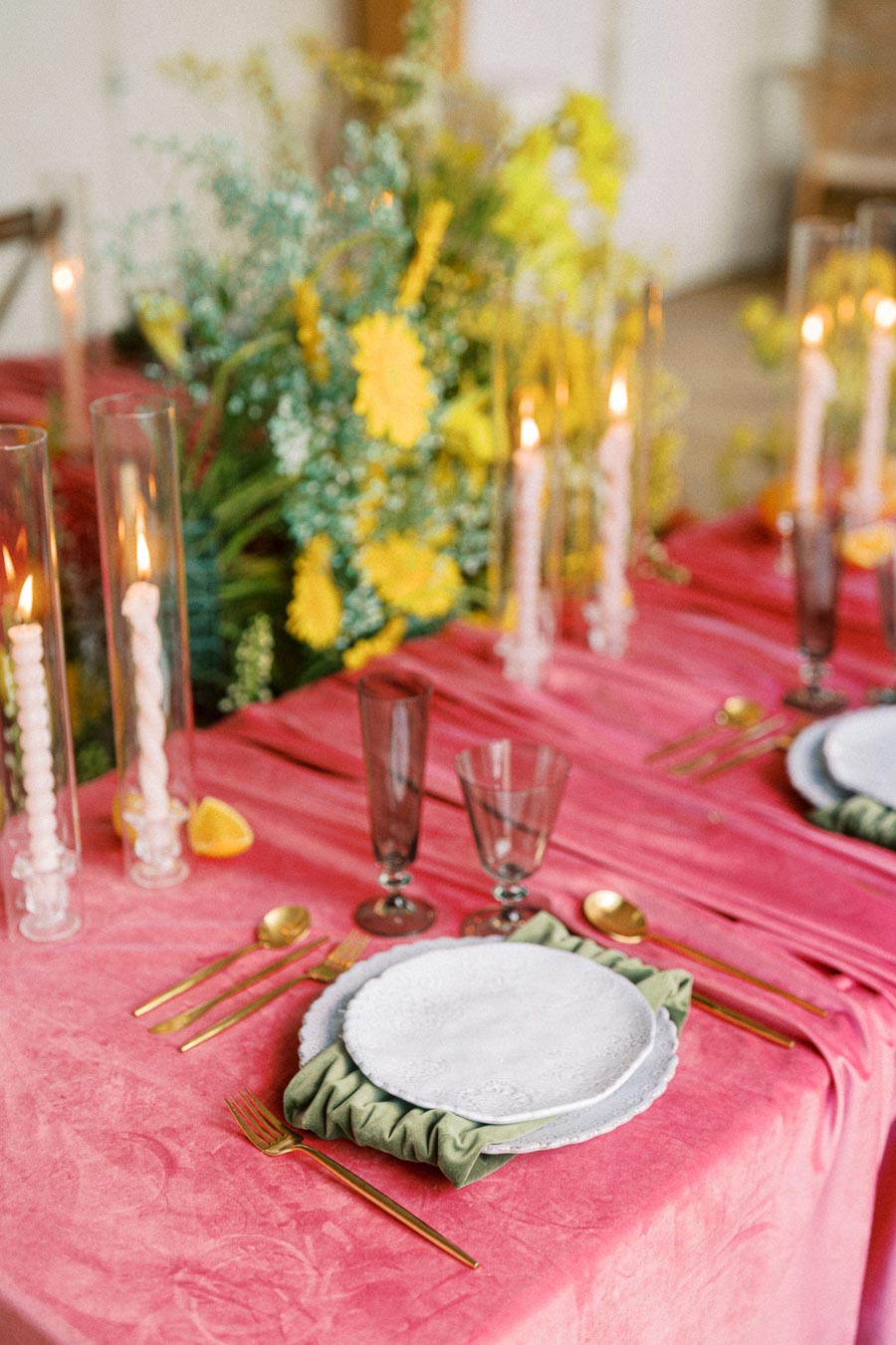 Elegant dining table setup with a vibrant pink tablecloth, adorned with gold cutlery, white textured plates on a green napkin, tall lit candles in glass holders, and a floral centerpiece featuring yellow and blue flowers.