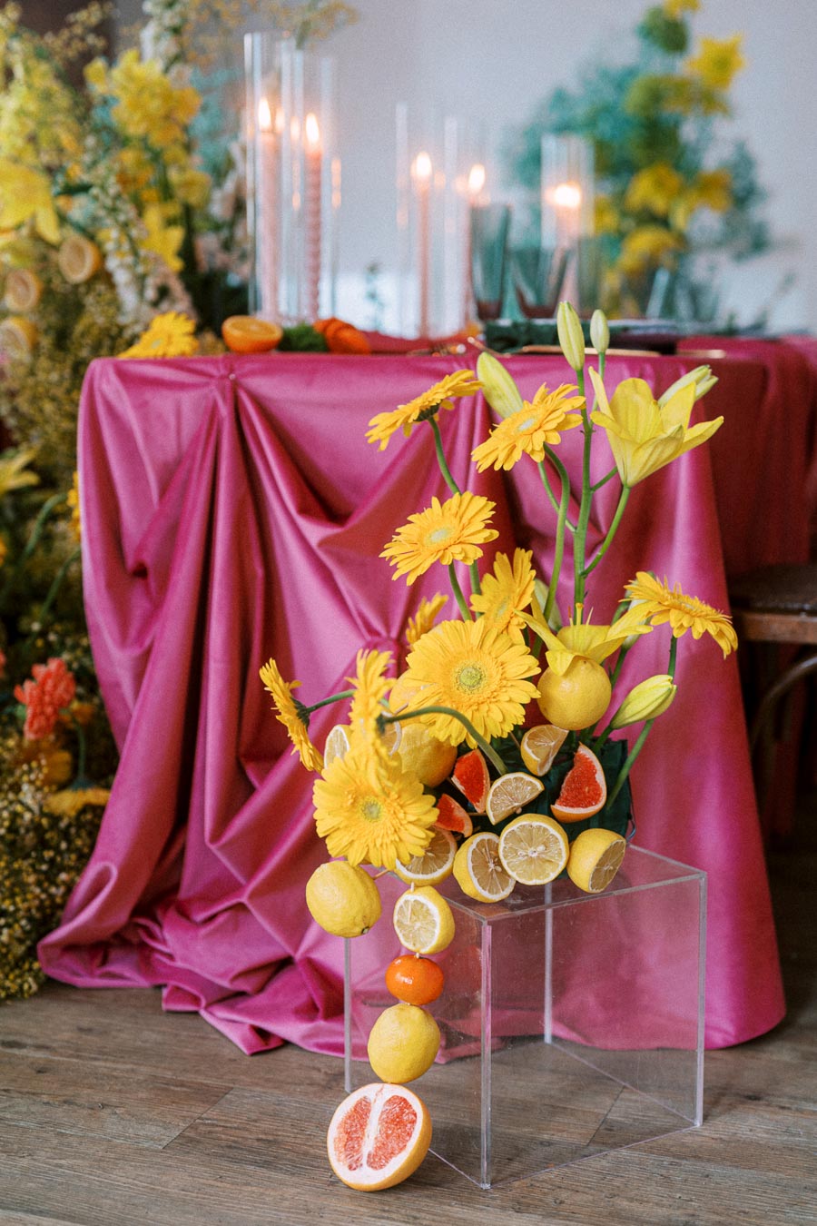 Yellow flowers and sliced citrus fruits arranged on a clear stand with a vibrant pink tablecloth backdrop, creating an elegant and colorful table setting accented by candles and greenery.