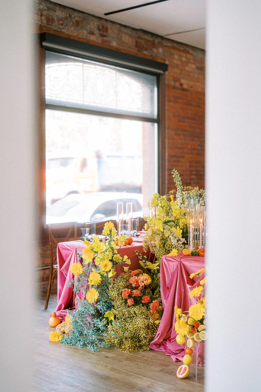 Elegant indoor floral arrangement with vibrant yellow and orange flowers on pink draped tables, framed by a large window and exposed brick wall, creating a charming, rustic ambiance.
