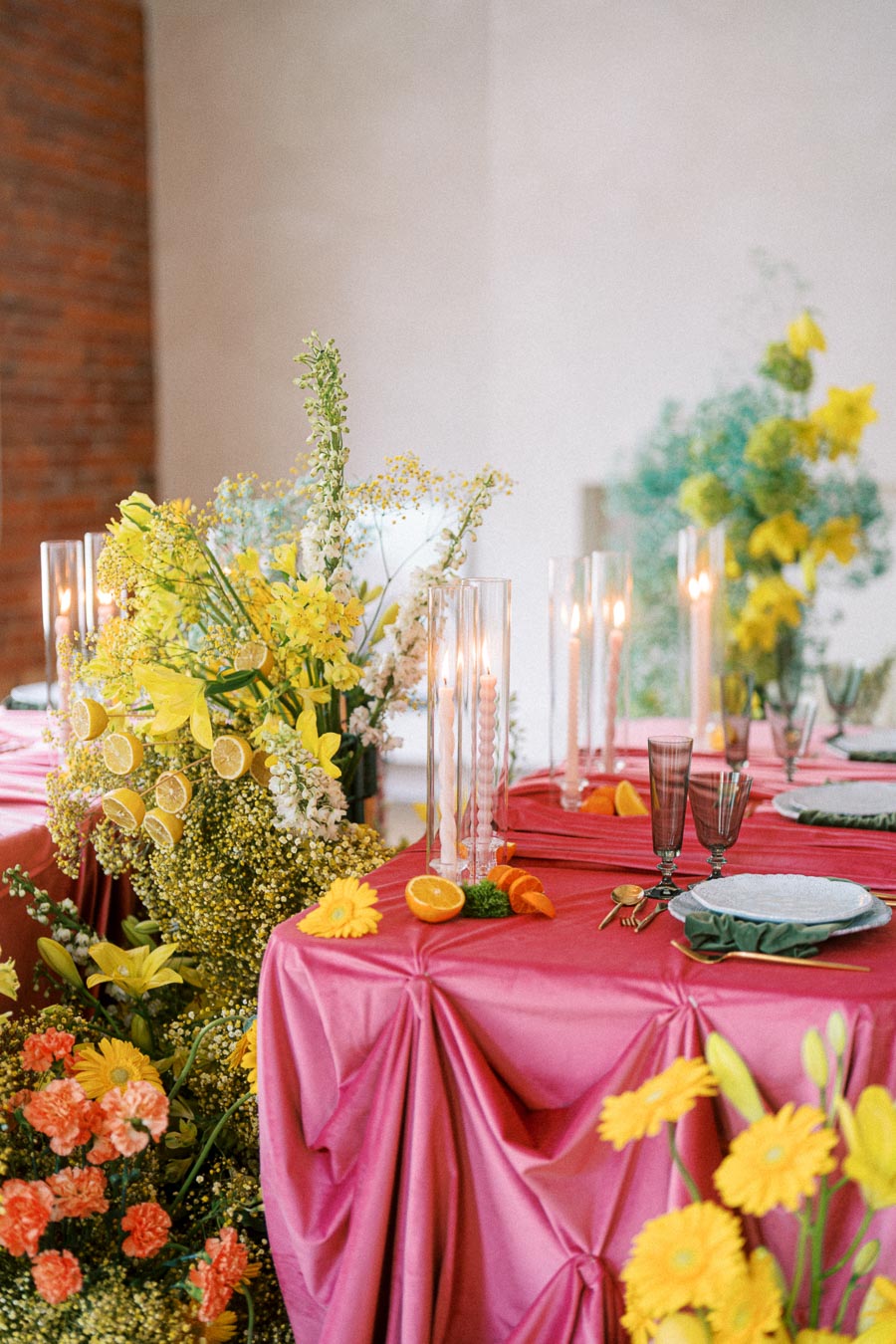 Elegant table setting with pink tablecloths, vibrant yellow and orange floral centerpieces, and slices of citrus fruits. Tall glass candles add sophistication, enhancing the festive, stylish atmosphere. Perfect for weddings or special events.