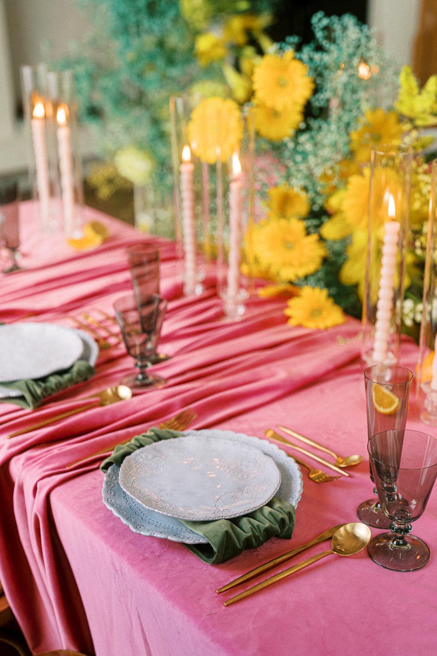 Elegant table setting with vibrant pink tablecloth, gold cutlery, and decorative plates, accented by tall candles and bright yellow flowers, creating a lively and stylish dining atmosphere.