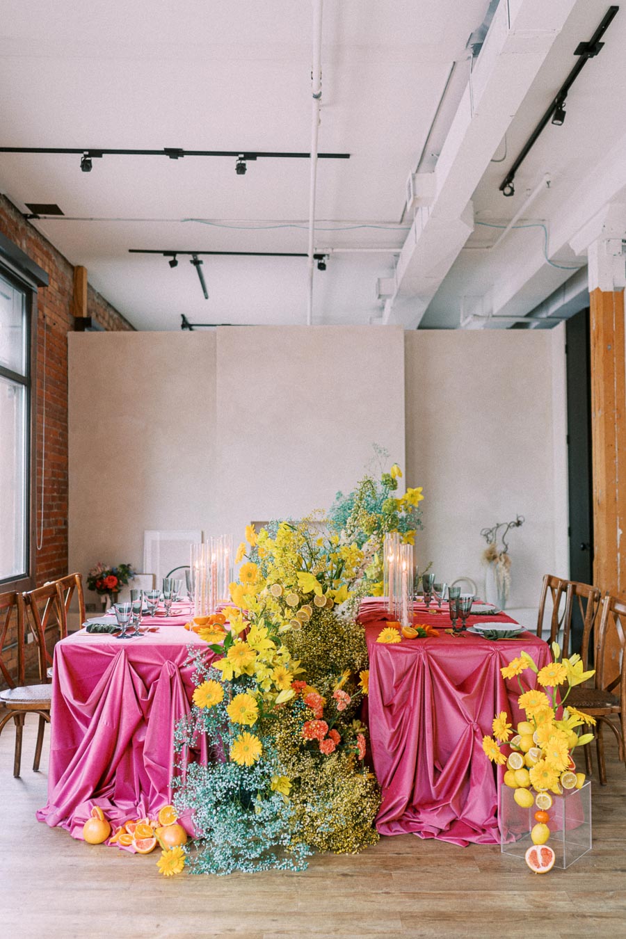 Elegant indoor dining setup with vibrant pink tablecloths, surrounded by yellow and orange floral arrangements, including daisies and roses. Glass vases adorn the table, which is set with elegant tableware and flanked by wooden chairs, creating a modern, colorful event decor.