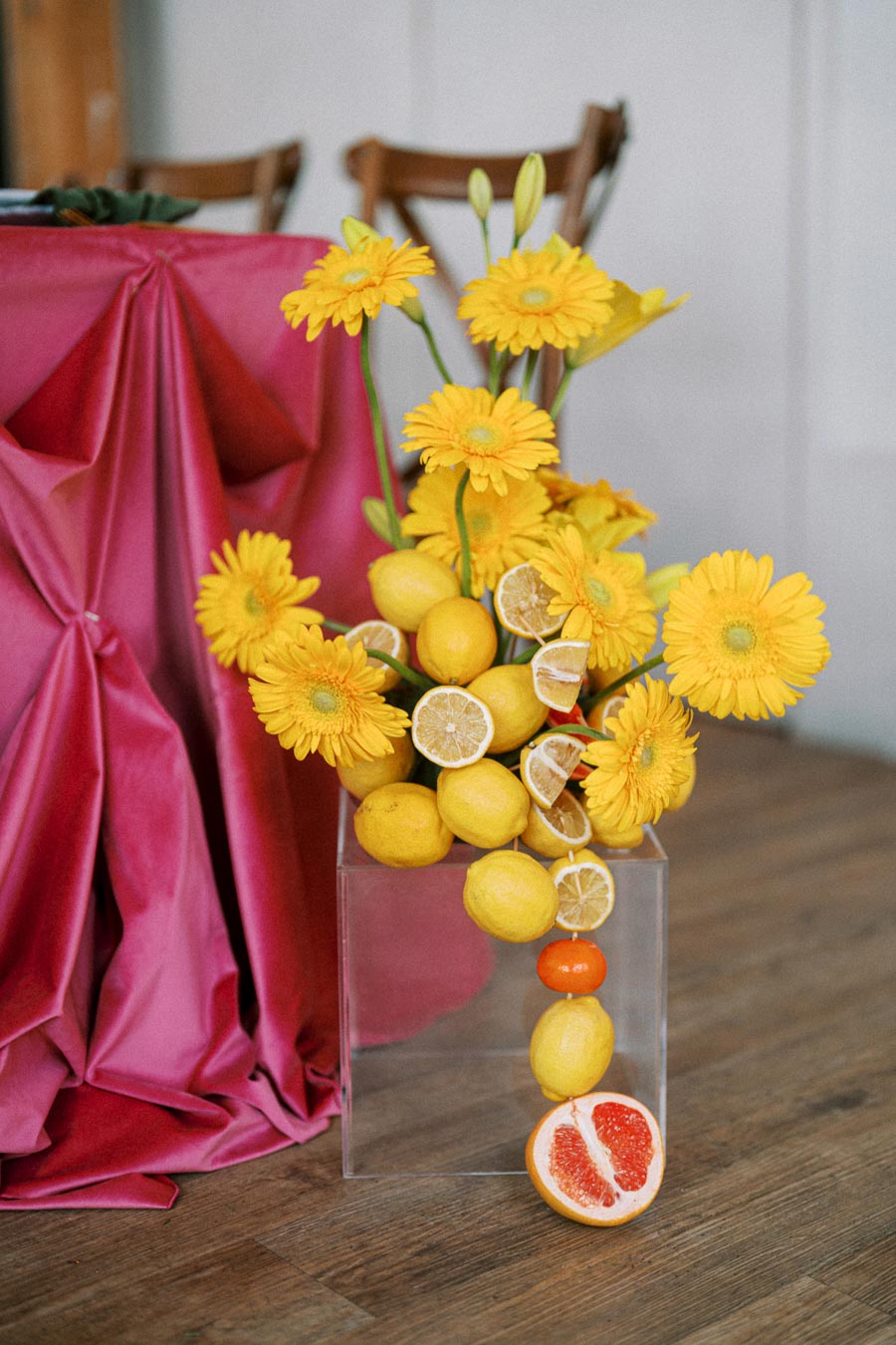 A vibrant floral arrangement featuring yellow gerbera daisies and lemons, complemented by sliced citrus fruits, artistically displayed in a clear vase. A rich pink drape and wooden chairs add a touch of elegance in the background. Perfect for a summer event or wedding decor.