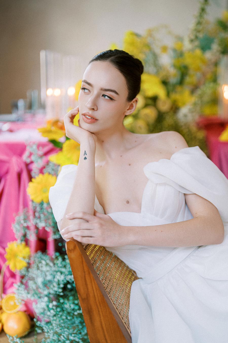 A woman in an elegant white off-shoulder dress poses thoughtfully on a wooden chair. The background is adorned with colorful flowers and lit candles, creating a vibrant and serene atmosphere.