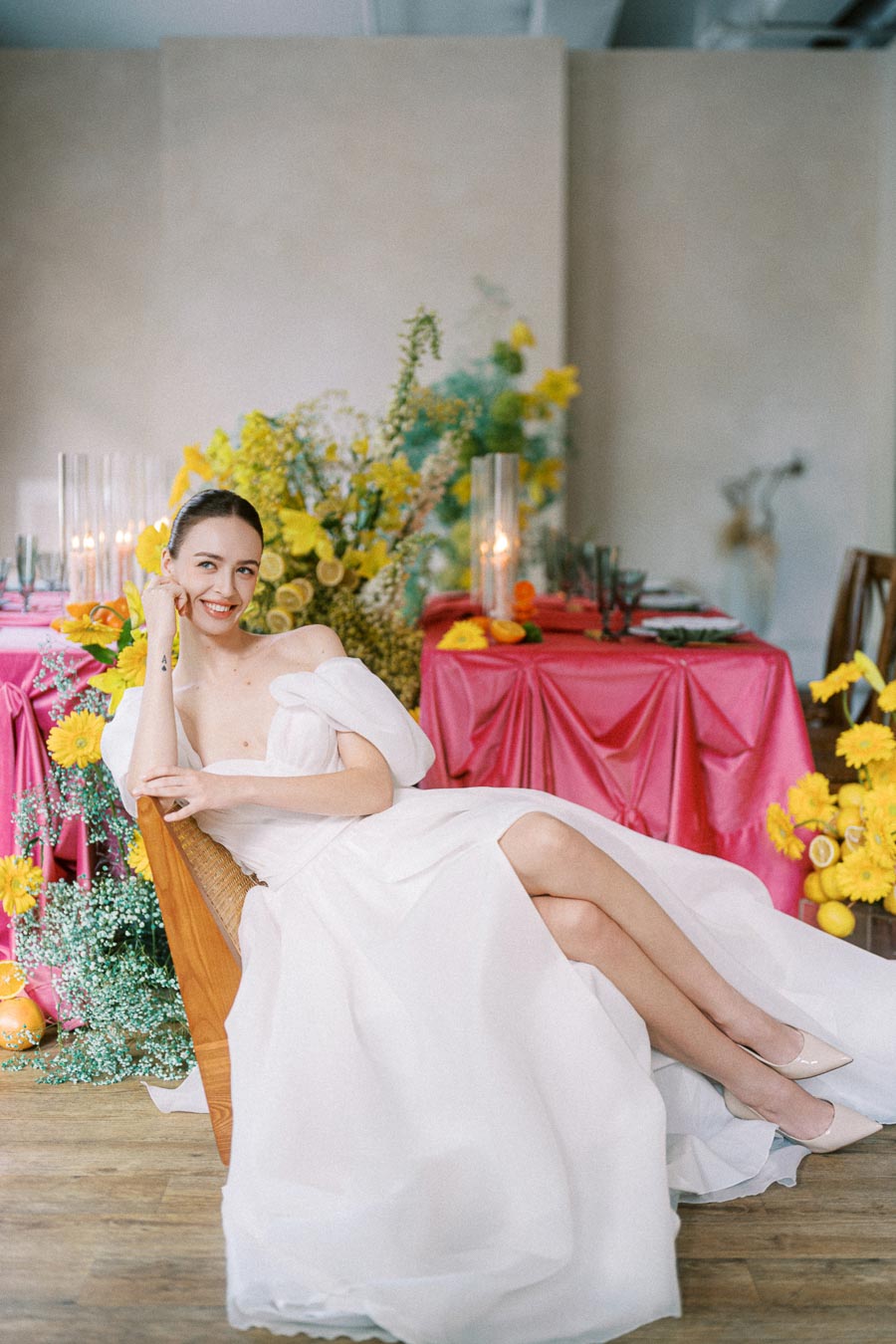 A woman in an elegant white dress sits on a wooden chair, smiling at the camera. The background features a vibrant setting with yellow flowers and a table draped in pink fabric, creating a cheerful ambiance.