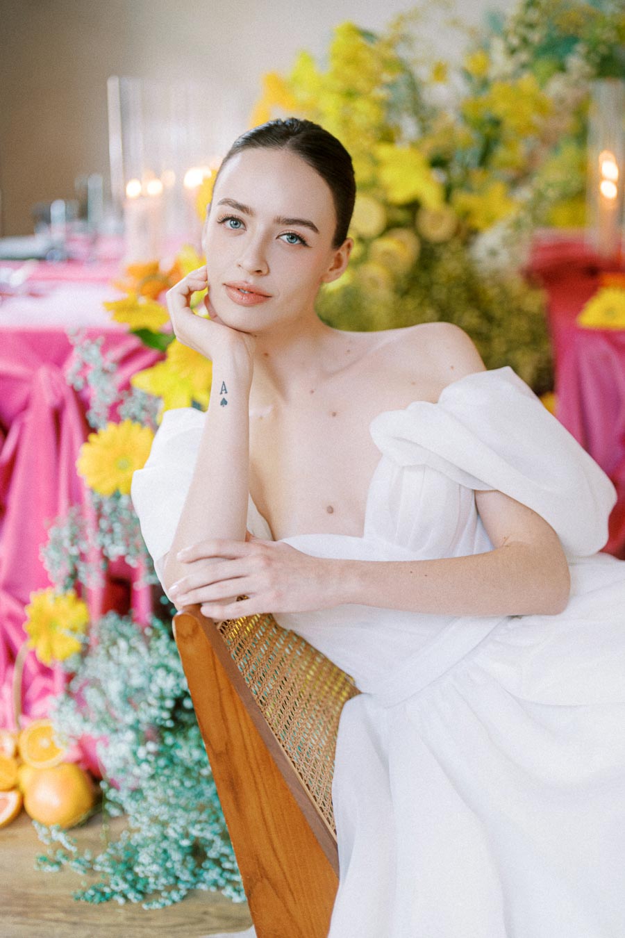 A woman in an elegant white off-shoulder gown poses seated, surrounded by vibrant yellow and pink floral arrangements, creating a colorful and stylish atmosphere.