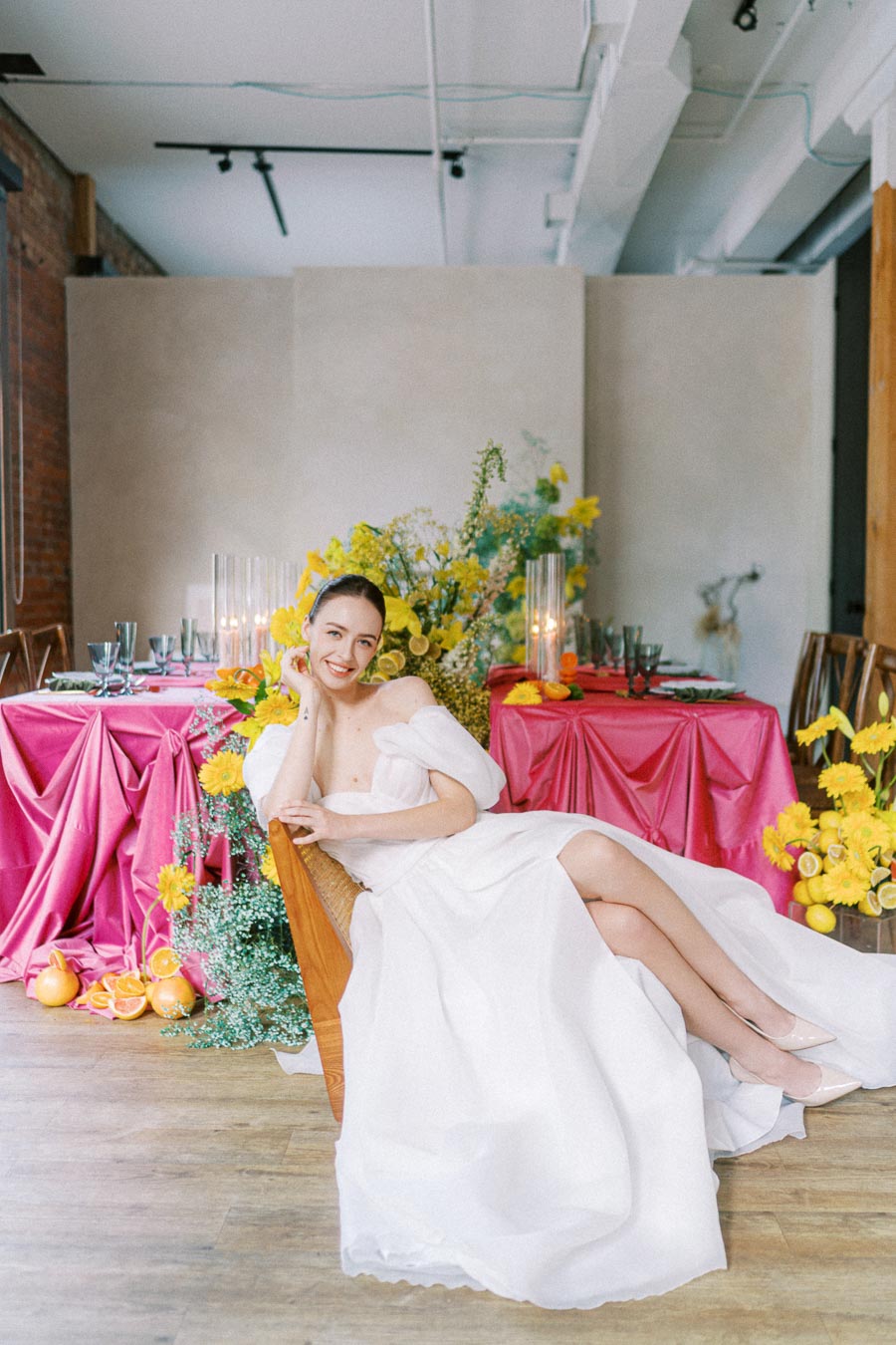 Bride in an elegant white dress smiling at a vibrant, colorful wedding setup with pink tablecloths, yellow flowers, and decorative citrus accents.