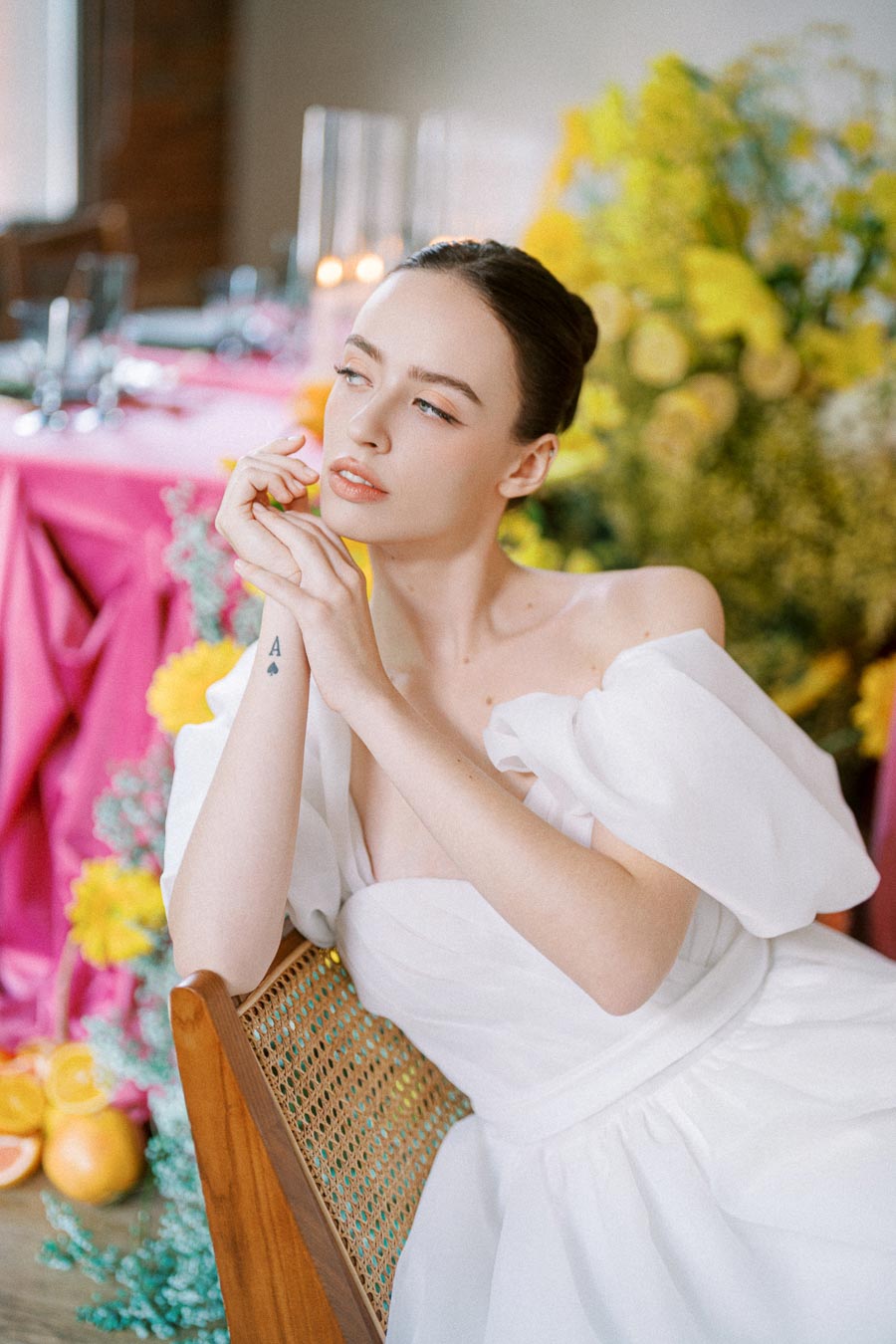 Young woman in an elegant white off-shoulder dress with a small ace of spades tattoo on her wrist, sitting on a wooden chair. The background features vibrant flowers, including yellow and pink blooms, and a beautifully set table with pink accents, creating a romantic and stylish atmosphere.