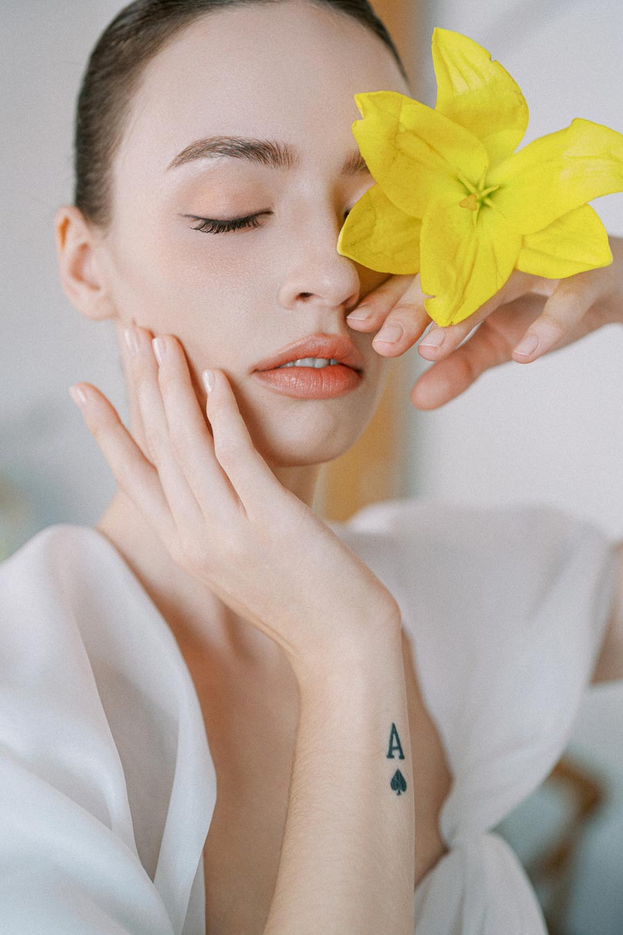 A woman with a serene expression holds a vibrant yellow flower near her face, showcasing her smooth skin and delicate tattoo of an ace of spades on her wrist. The soft lighting and close-up angle emphasize her natural beauty and tranquil atmosphere.
