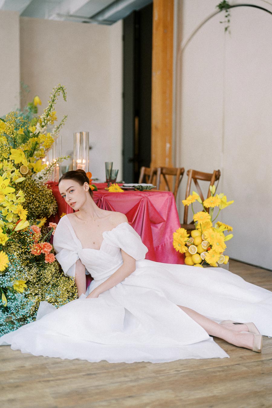 A woman in an elegant white dress poses beside a vibrant floral arrangement and a table set with a vivid pink tablecloth, featuring bright yellow and orange flowers, creating a stunning and colorful wedding or event scene.