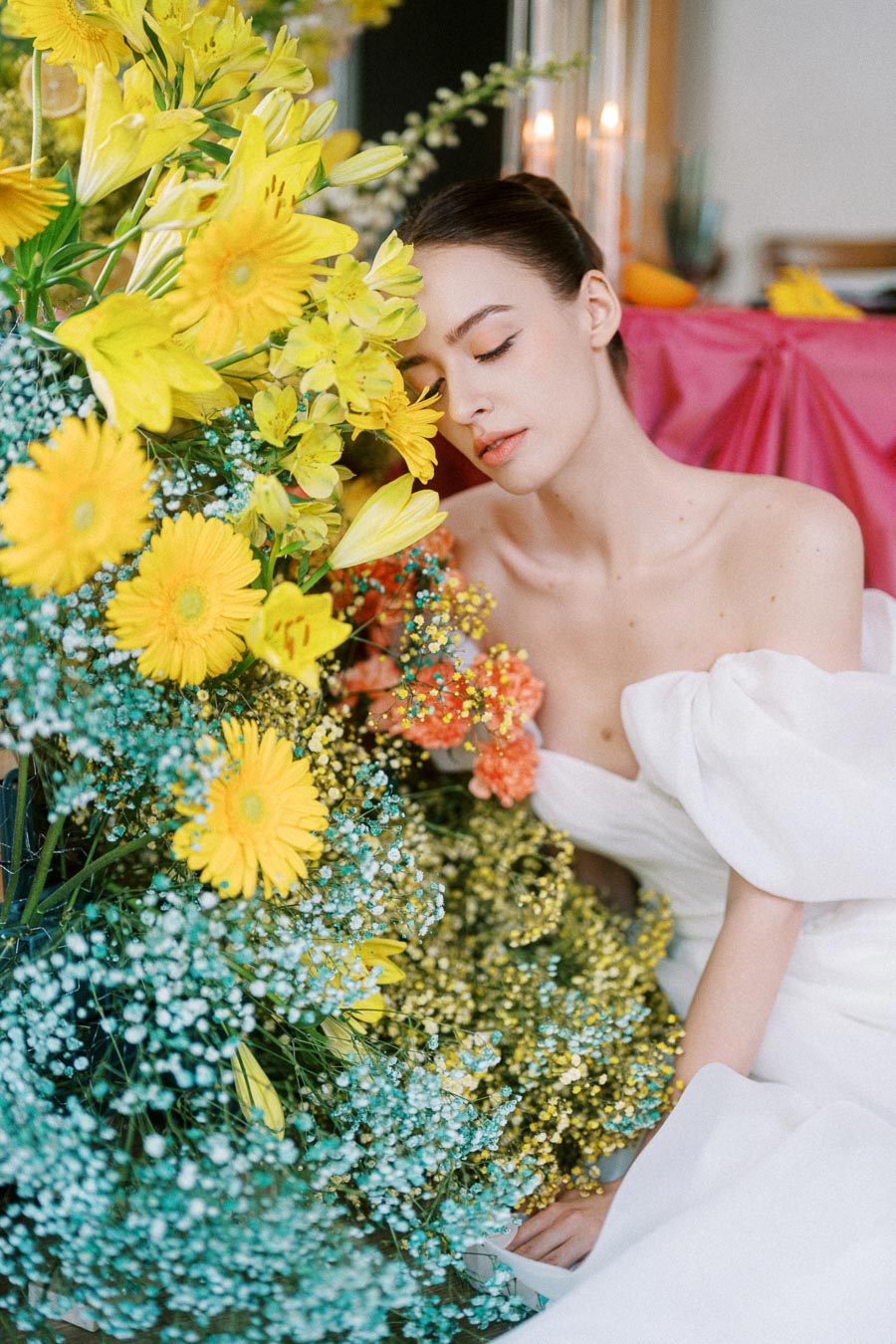 A woman in a white dress poses beside a vibrant bouquet of yellow flowers, including daisies and lilies, against a pink backdrop.