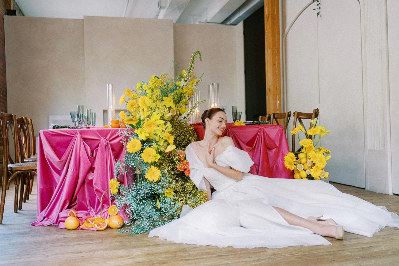 Elegant woman in a flowing white dress sitting beside a vibrant table setting with pink drapery and a lush arrangement of yellow flowers and citrus fruits.