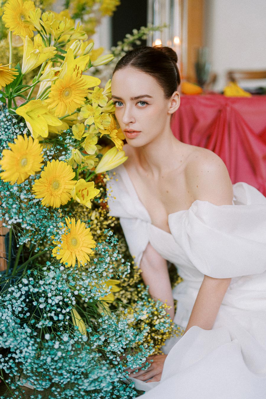 A woman in an elegant white gown sits beside a vibrant arrangement of yellow flowers and greenery. The background features a draped pink fabric, adding a touch of elegance to the composition.