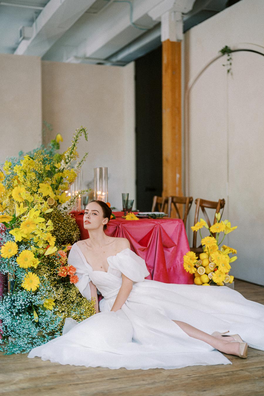 Bride in elegant white dress sitting next to vibrant floral arrangement with yellow flowers at a stylish indoor wedding venue, featuring a red tablecloth and modern decor.