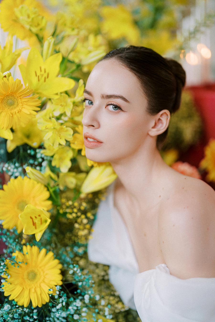 A woman surrounded by vibrant yellow flowers, including daisies and lilies, wearing a white off-shoulder top, exuding elegance and serenity.