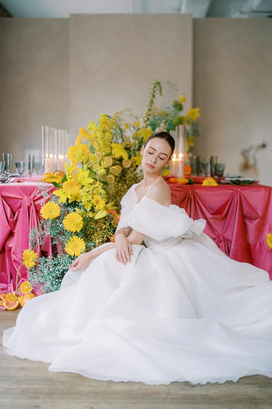 Elegant bride in a flowing white dress sitting by a vibrant floral arrangement with yellow flowers and oranges, next to a pink draped table setting, creating a romantic wedding scene.