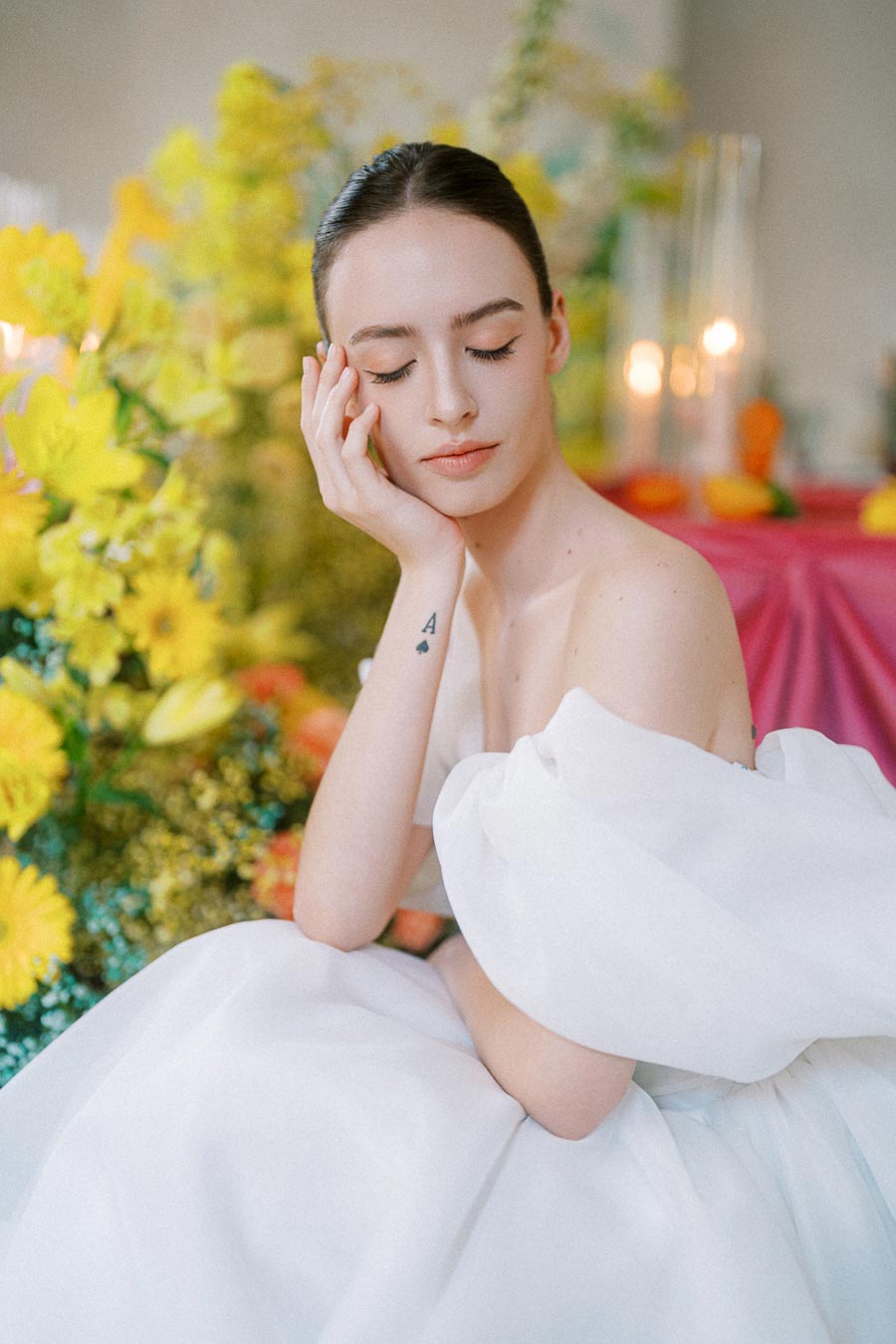 A woman in a white off-shoulder gown with a tattoo of an ace of spades on her arm poses gracefully against a backdrop of vibrant yellow flowers and softly lit candles.