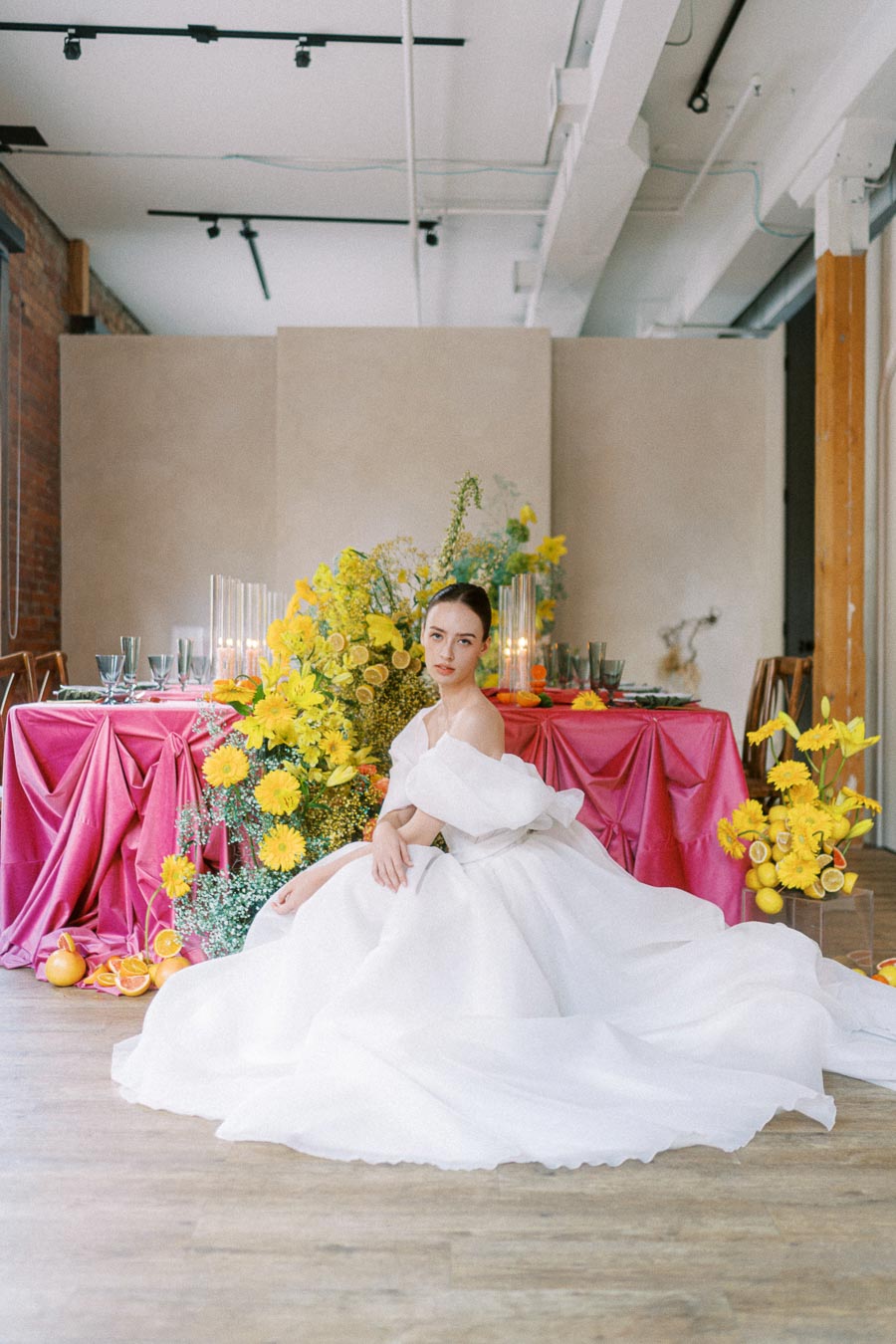 Elegant bridal portrait featuring a bride in a flowing white gown, seated in a modern industrial venue. The setting includes a vibrant pink tablecloth, vivid yellow floral arrangements, and soft candlelight, creating a romantic wedding atmosphere. Perfect inspiration for contemporary wedding decor ideas.