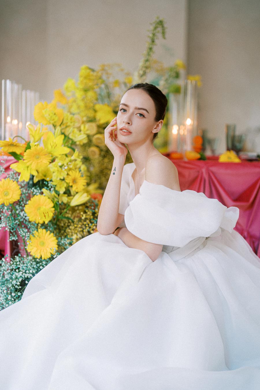 A woman in an elegant white gown poses thoughtfully amidst a vibrant floral arrangement of yellow flowers and greenery, creating a serene and artistic wedding scene.