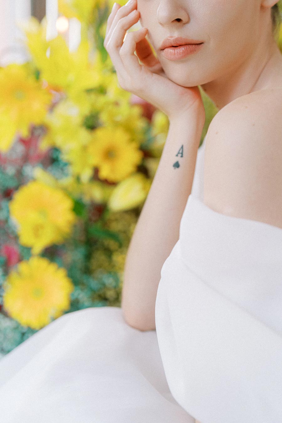 A woman in a white dress with a visible tattoo of an A and a spade on her arm, holding her face gently with her hand. She is surrounded by a vibrant bouquet of yellow flowers.