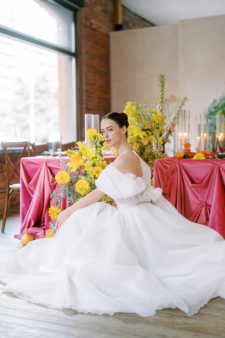 Bride in an elegant white wedding gown posing beside a vibrant, colorful table setting with yellow flowers and pink tablecloth, creating a sophisticated and romantic atmosphere.