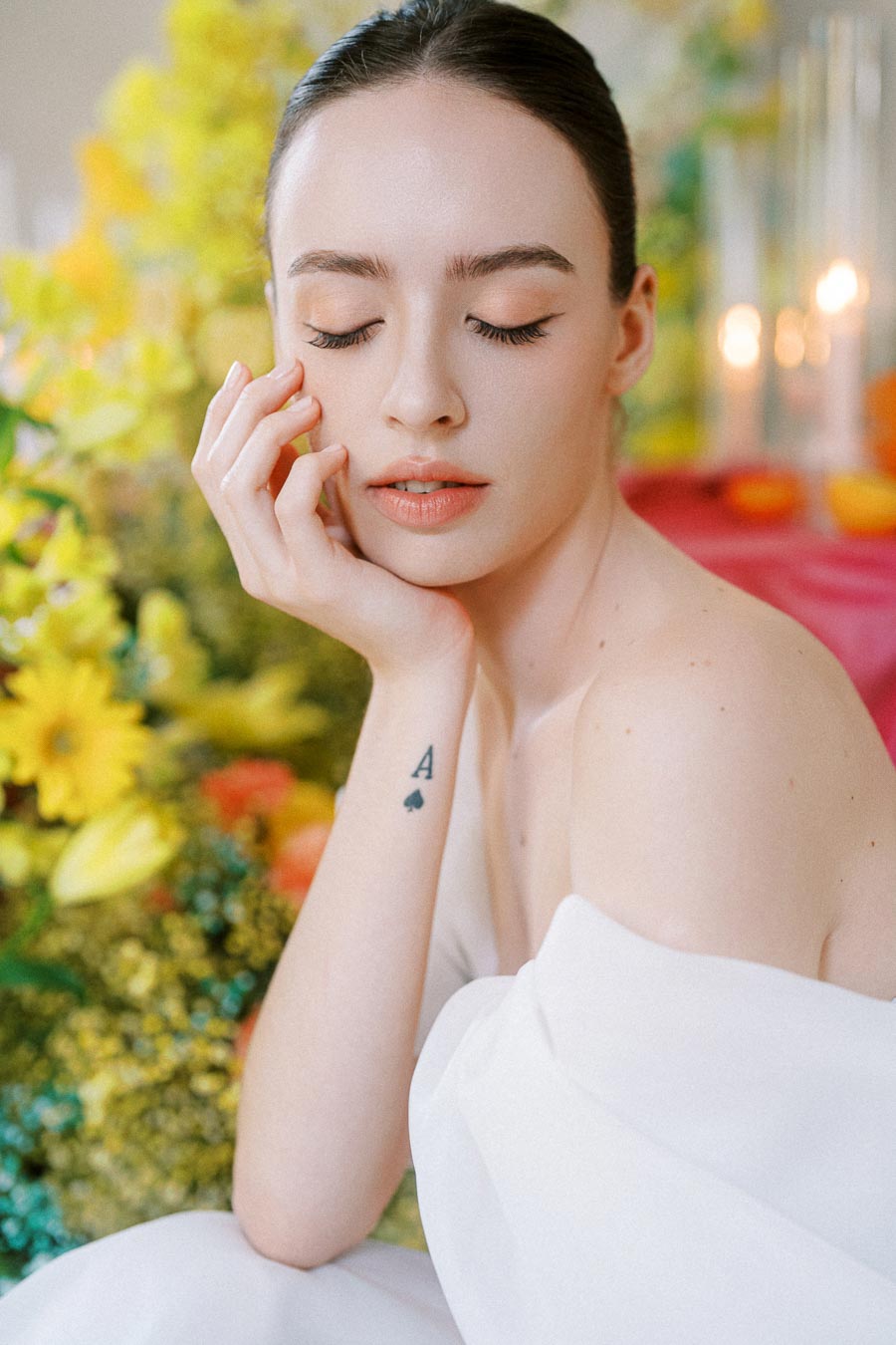 A serene woman in a white dress with eyes closed, surrounded by vibrant yellow flowers and soft candlelight, displaying an ace of spades tattoo on her arm.