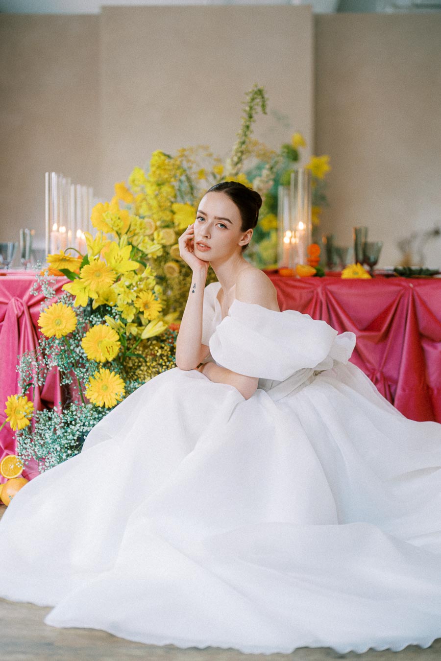 Bride in elegant white gown sitting by a vibrant floral arrangement with yellow flowers and a pink draped table, creating a striking contrast in a wedding setting.