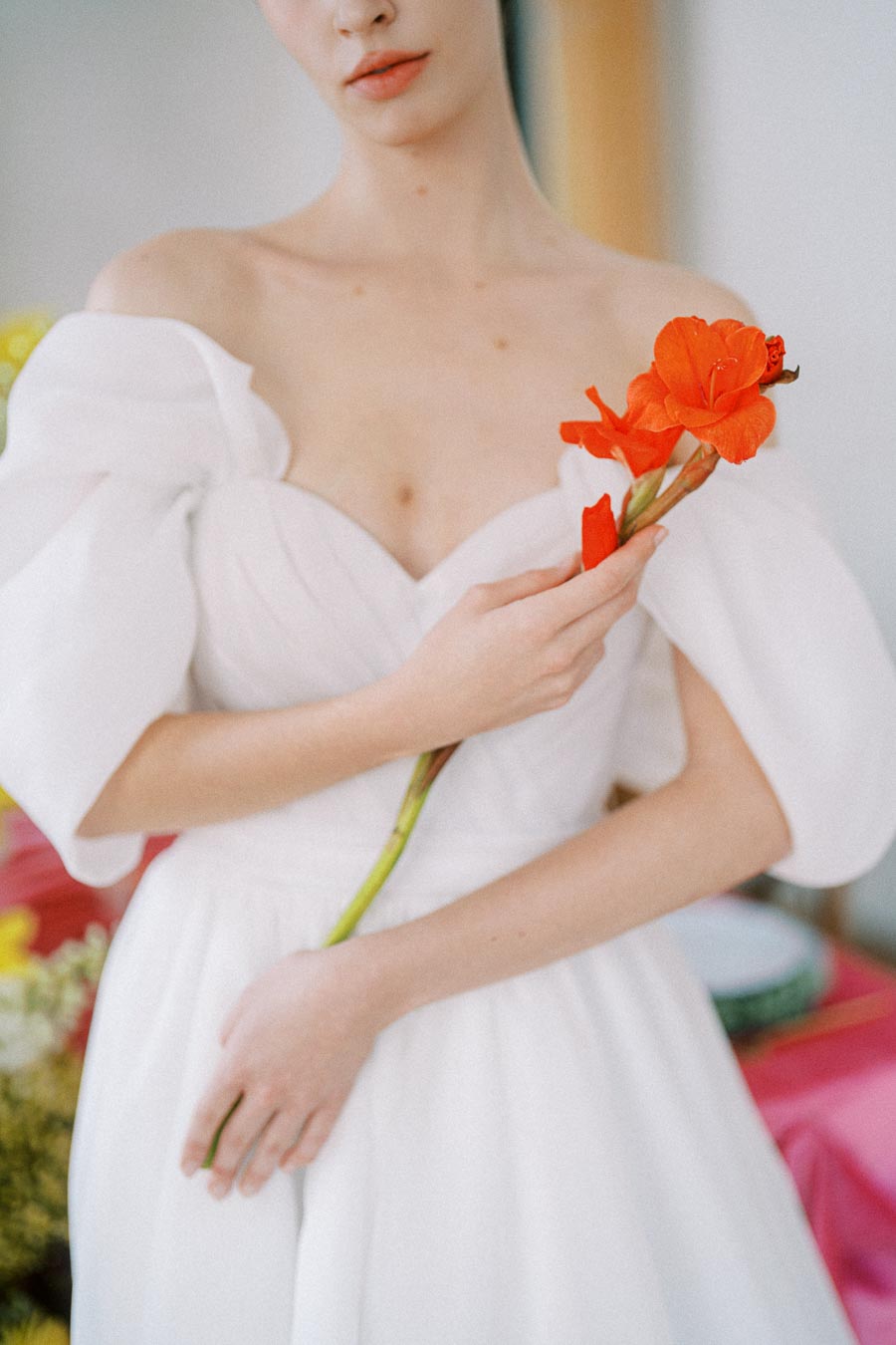 A woman in an elegant white dress holds a vibrant orange flower, showcasing a blend of natural beauty and sophisticated fashion.