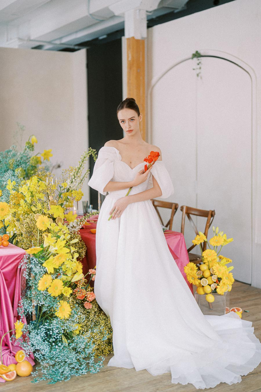 Elegant bride in a flowing white wedding gown holding a red flower, standing beside a vibrant floral arrangement with yellow blooms and citrus accents in a tastefully decorated indoor setting.