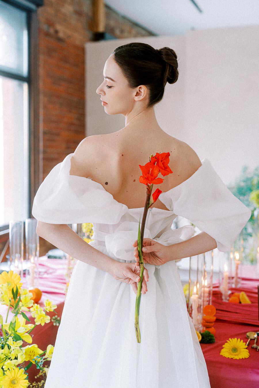 Elegant bride holding a vibrant red flower behind her back, dressed in a white off-shoulder gown, with a beautifully set table with yellow flowers in the background.