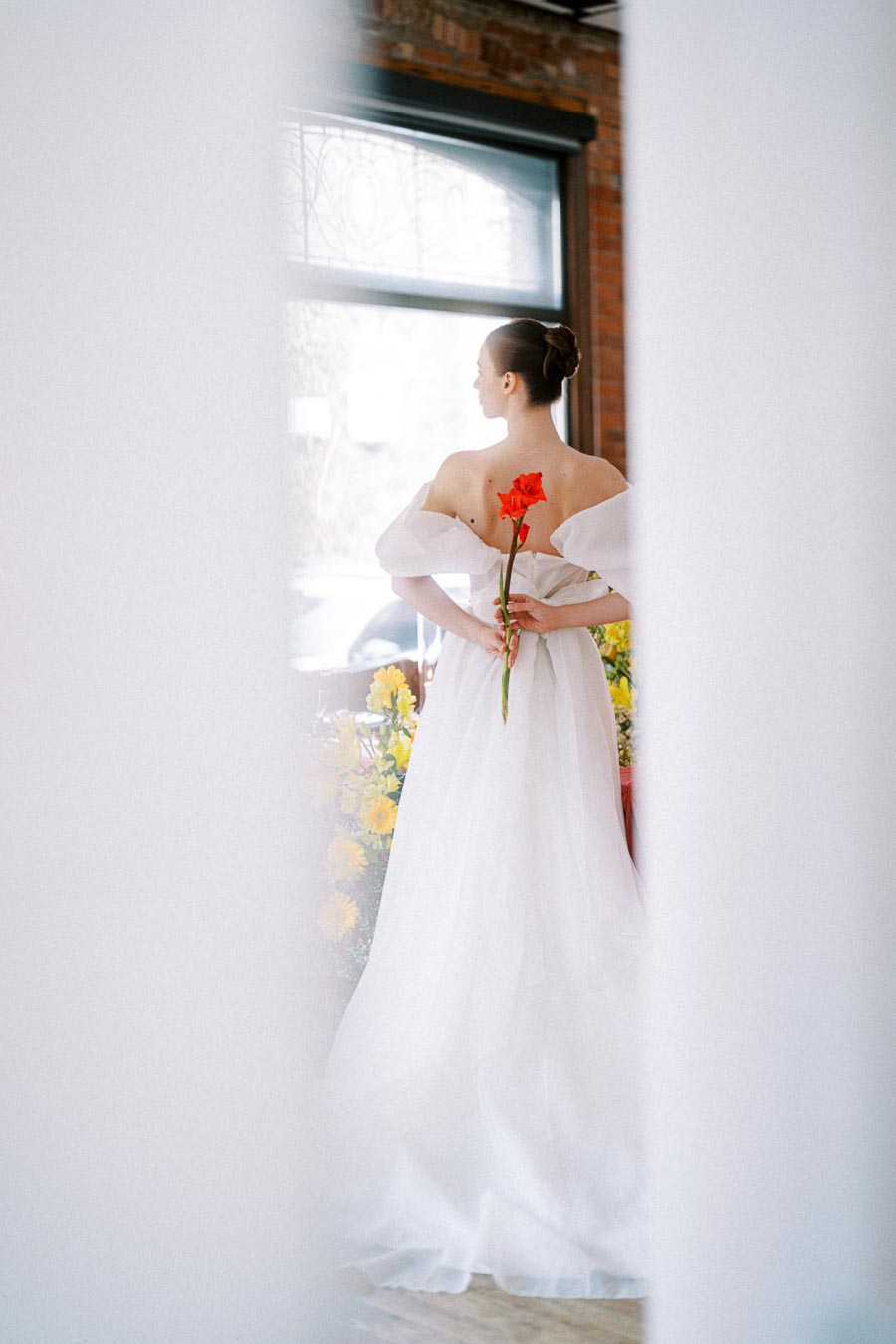 Elegant bride in white gown holding a vibrant red flower, standing in a bright room with floral decorations around.