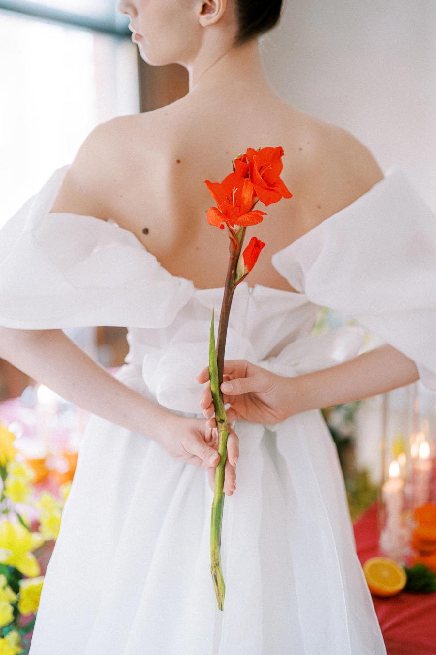 Elegant woman in white dress holding a vibrant red gladiolus flower behind her back, surrounded by soft lighting and floral decor.