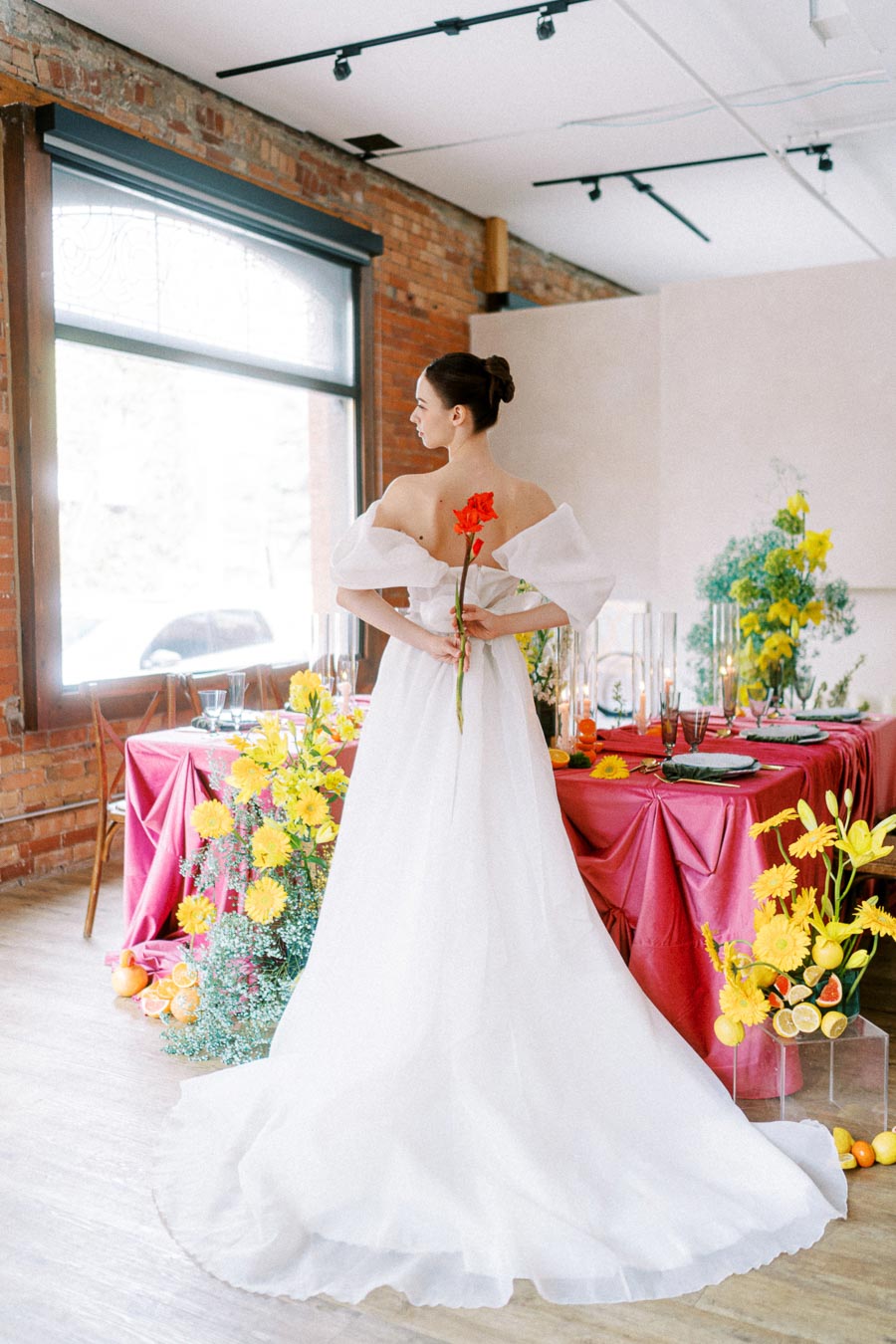 Bride in elegant white gown holding red flower, standing in vibrant floral-themed wedding venue with pink table setting and yellow floral arrangements.