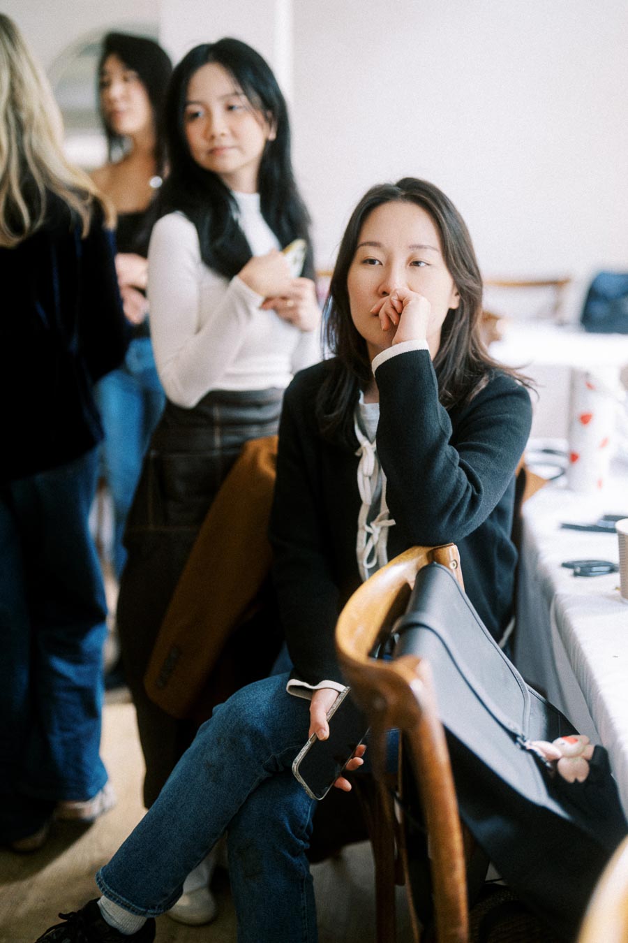 Group of women in a casual social setting, with one seated and holding a phone, engaged in conversation.