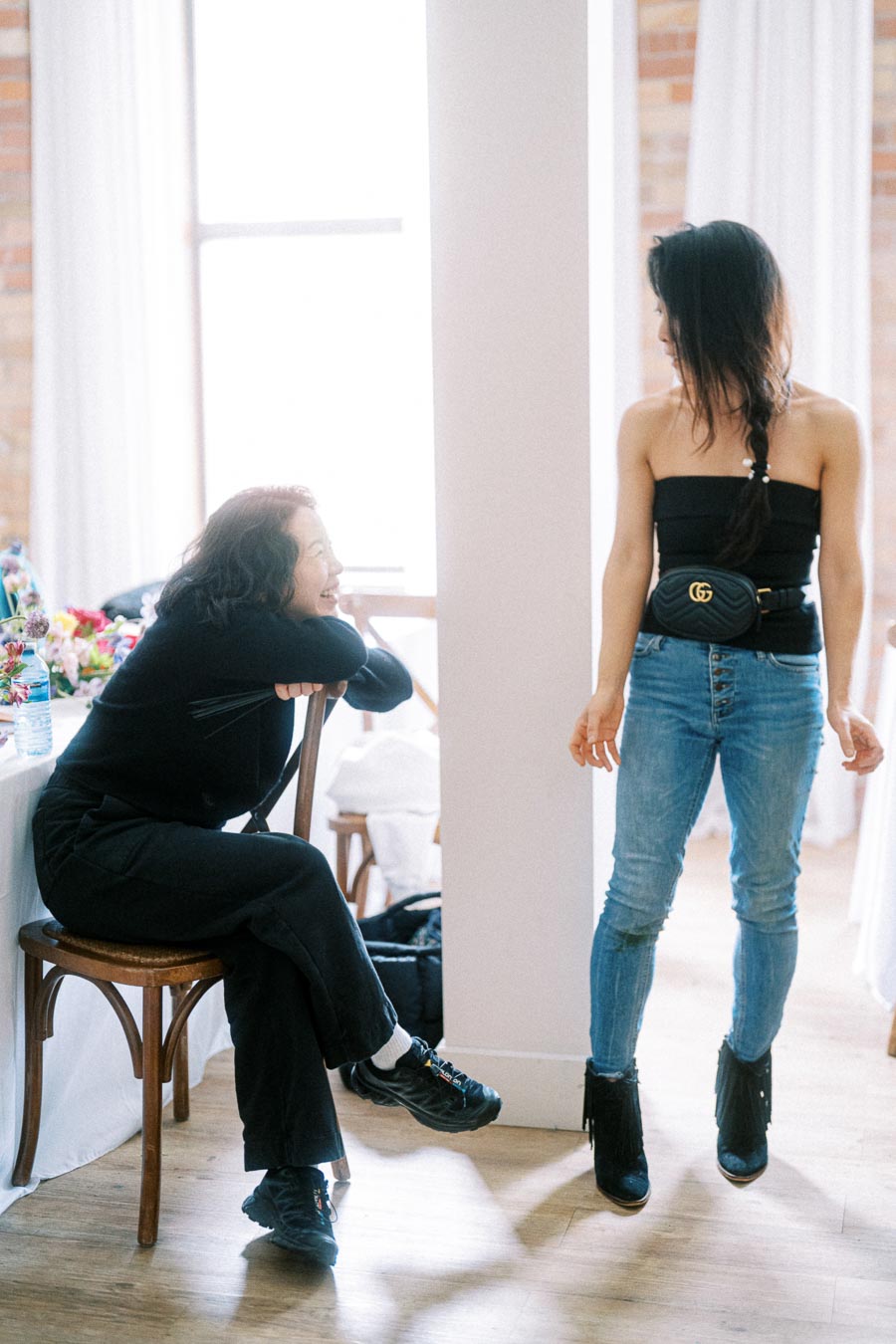 Two women in casual conversation indoors. One is seated on a chair, smiling and leaning on the table, while the other stands facing her with a braided hairstyle and wearing jeans with a black top. Sunlight filters through a large window, highlighting the relaxed and cheerful atmosphere.