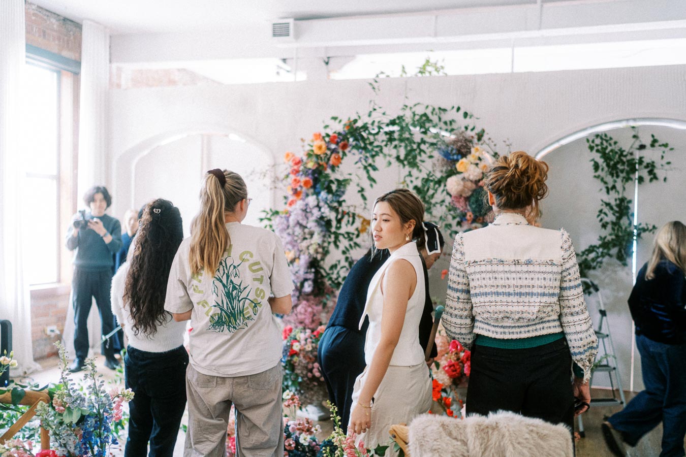 Group of people in a bright room with floral decorations, featuring colorful flowers and greenery, engaged in an event or discussion.