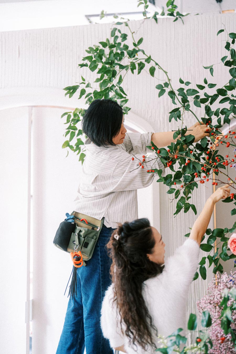 Florists arranging decorative greenery and berries on a white archway for an event setup, highlighting teamwork and creativity in floral design.