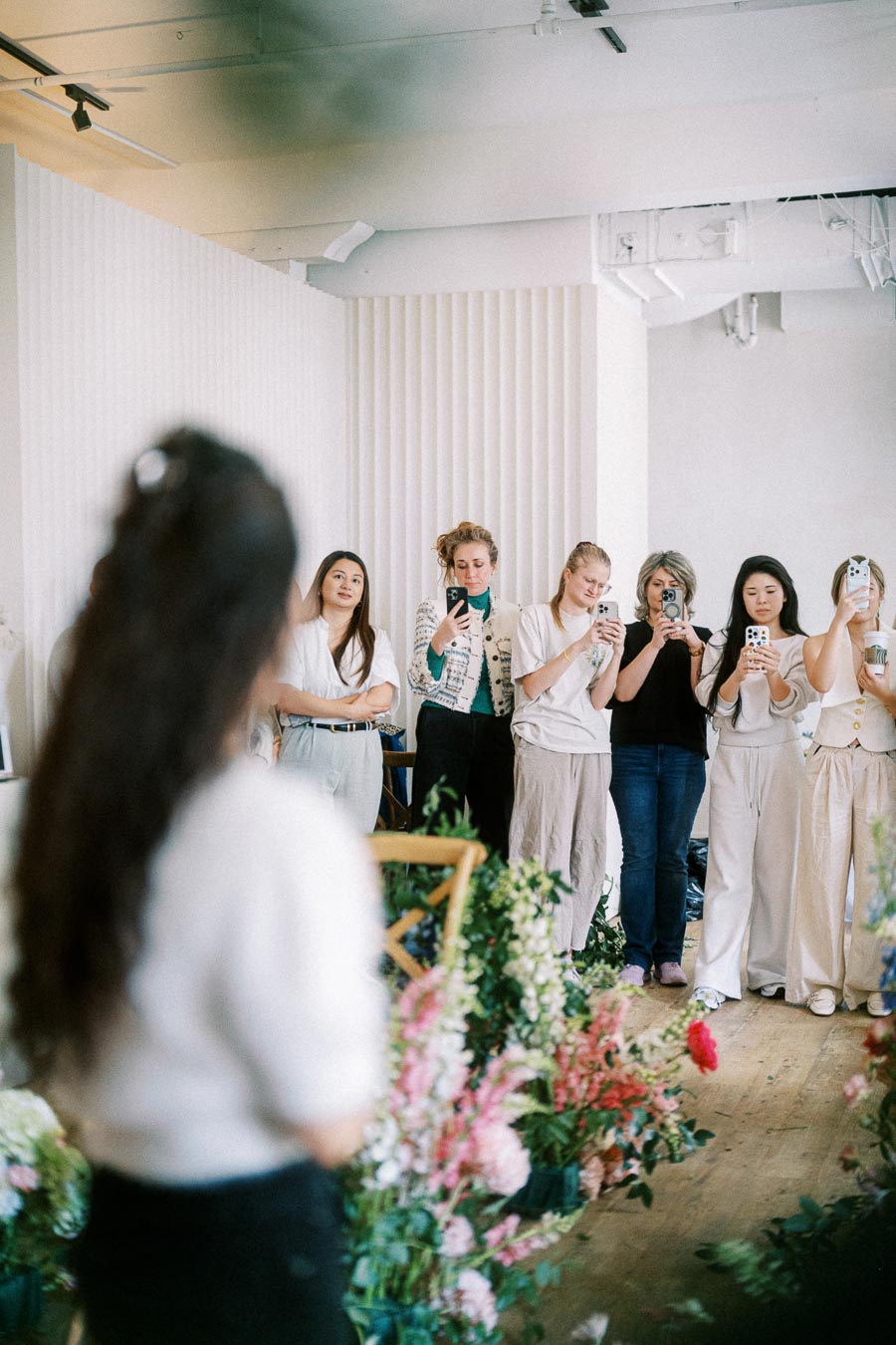 Group of people enthusiastically photographing a flower arrangement demonstration in a bright, modern room.