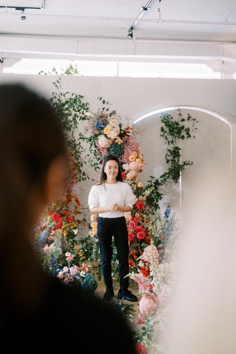 A person standing in front of a large, colorful flower arrangement indoors. They are wearing black pants and a white sweater, with a thoughtful expression. The floral display includes red, pink, and yellow blooms, creating a vibrant background. Bright indoor lighting highlights the scene.