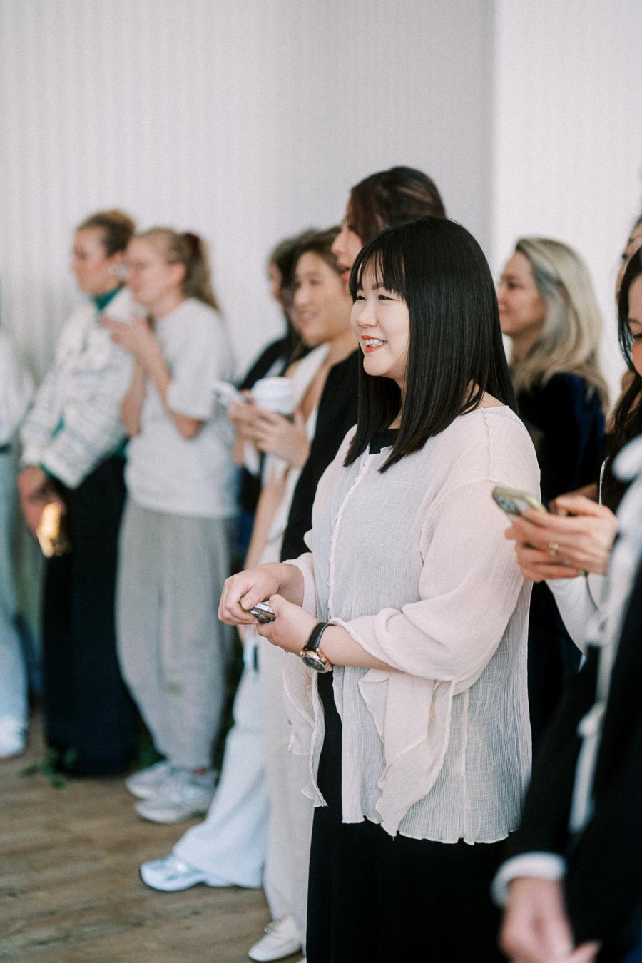 A group of diverse women standing in a room, engaged in conversation and smiling, some holding smartphones or cups, during a social event or networking gathering.